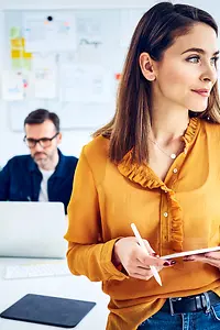 Businesswoman standing in a modern office with tablet and pen in hands, her colleague sitting in the background at laptop